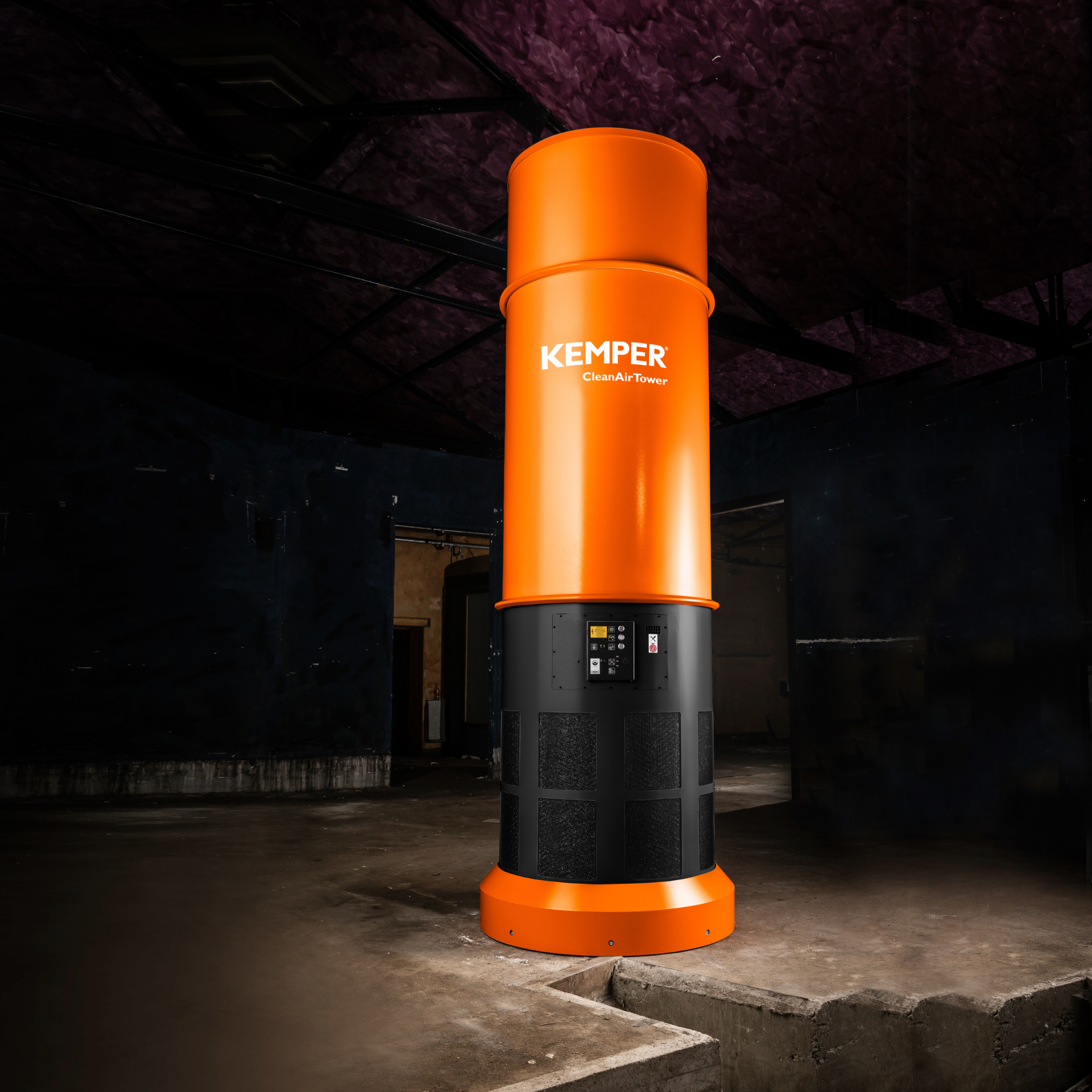 Freestanding cylindrical KEMPER CleanAirTower in orange and black with integrated control unit, installed in a dark industrial hall with concrete floor and steel structure.