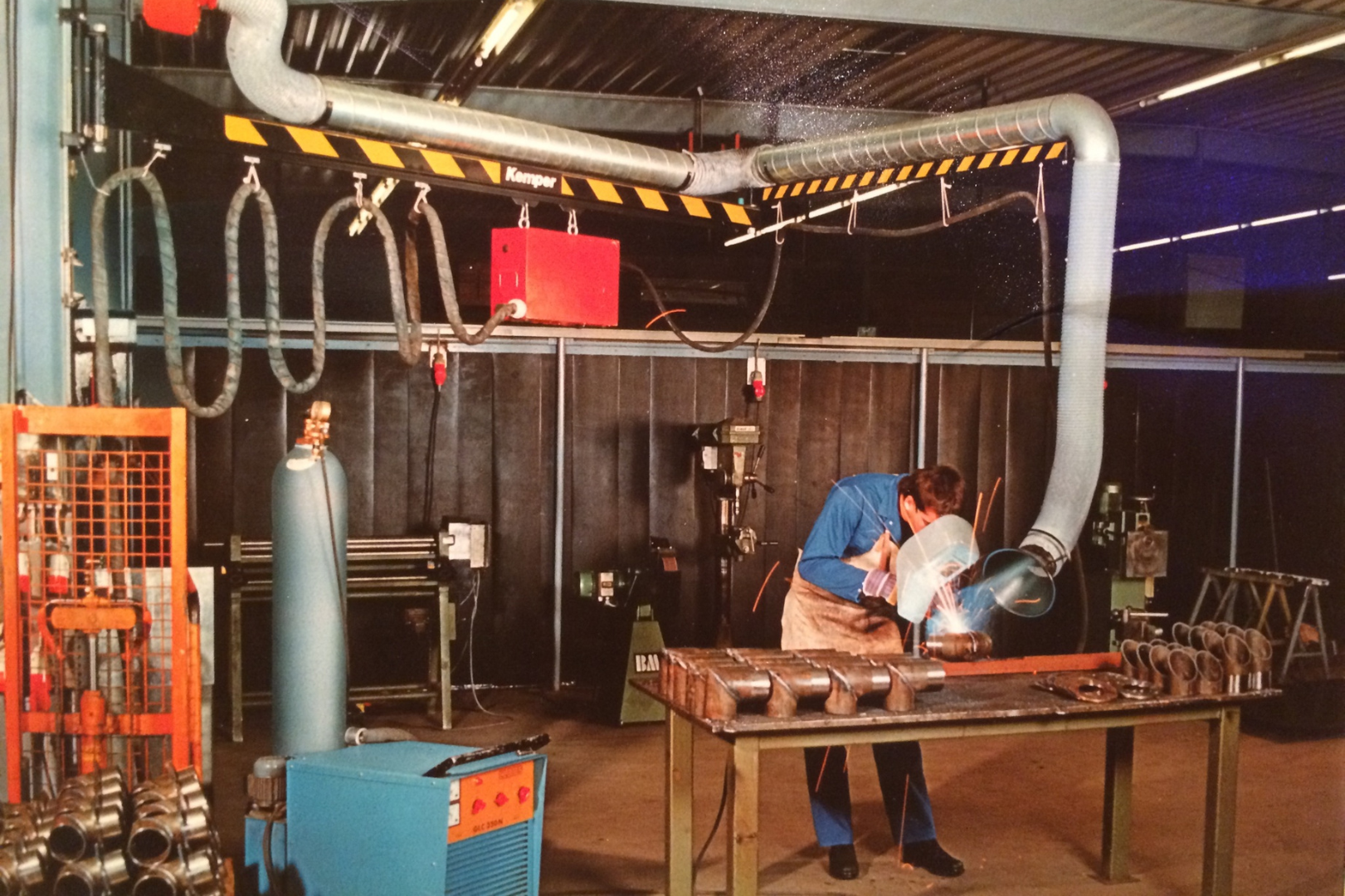 Worker welding metal parts in a workshop equipped with an extraction system and ducting for fume removal.