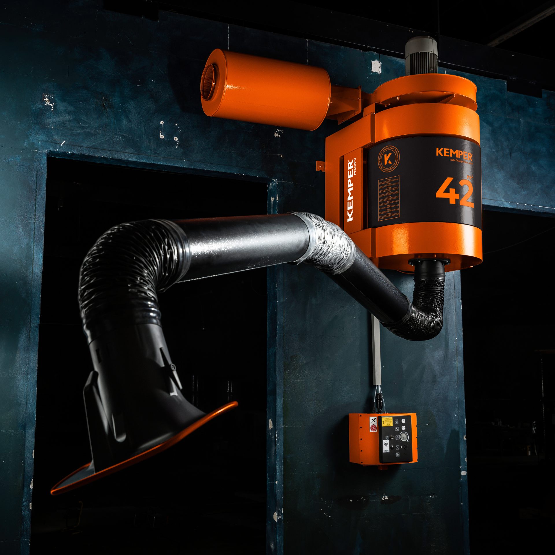 Orangefarbenes KEMPER WallMaster Absauggerät mit schwarzem, flexiblem Absaugarm und Filtereinheit (42 m²), montiert an einer industriellen Werkstattwand mit separatem Steuerkasten.