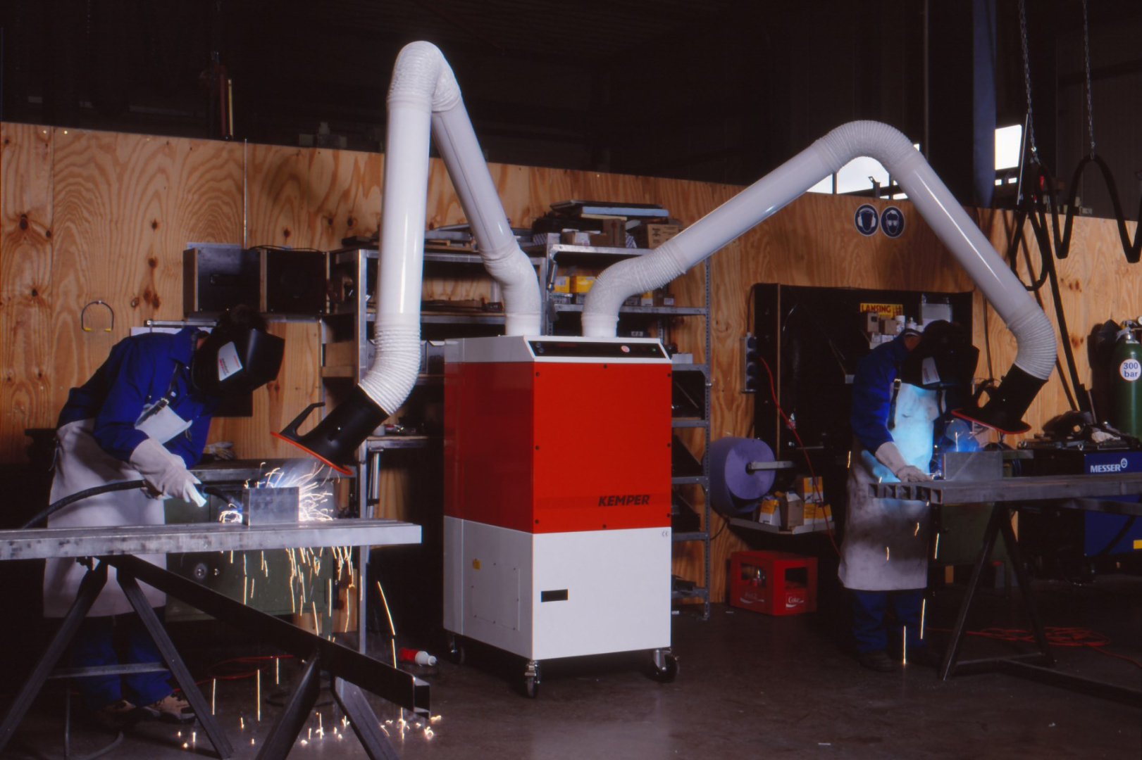 Two workers welding at workbenches equipped with a mobile extraction unit and flexible extraction arms for capturing fumes and sparks.