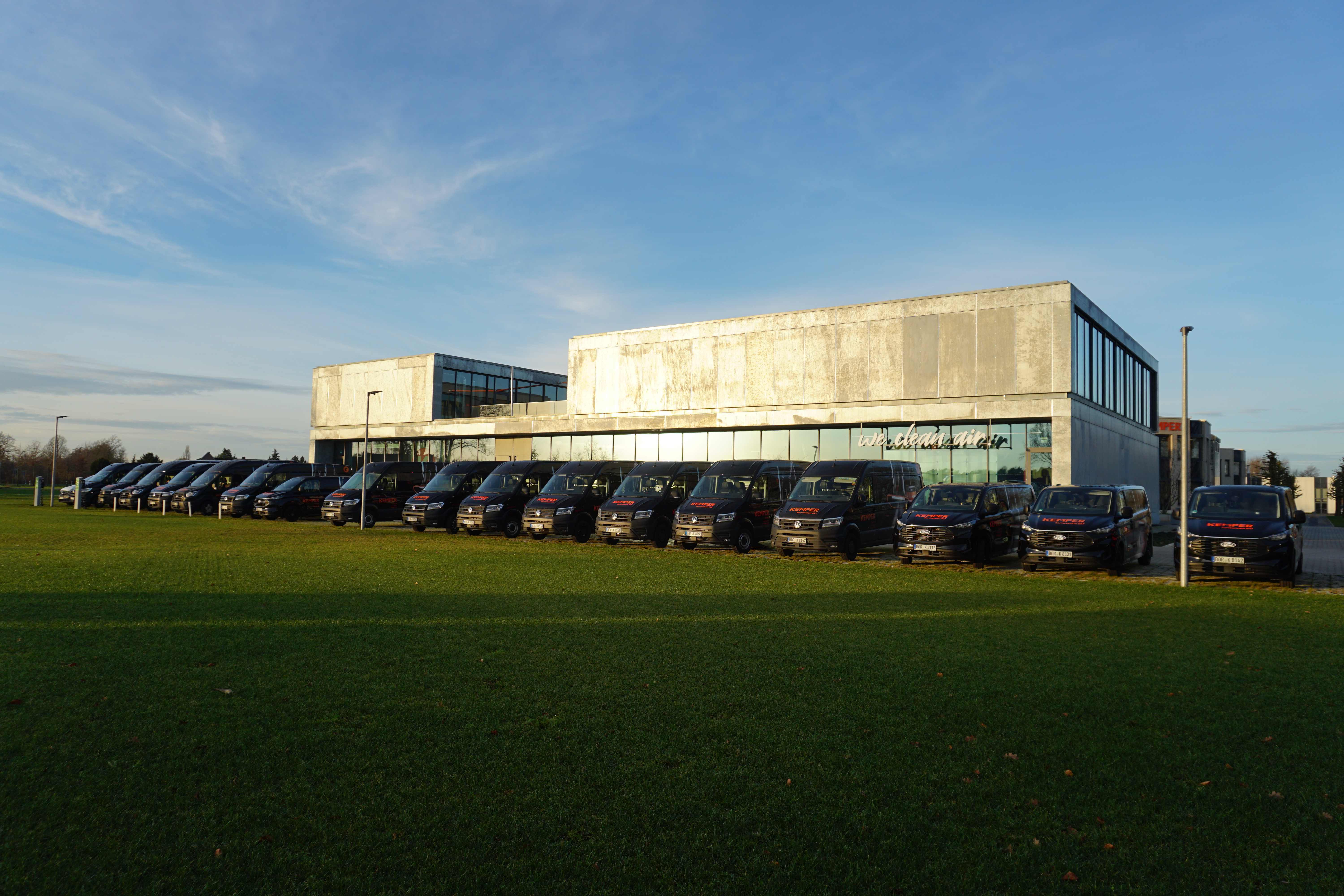 A row of KEMPER service vans parked in front of a modern company building on a green lawn.