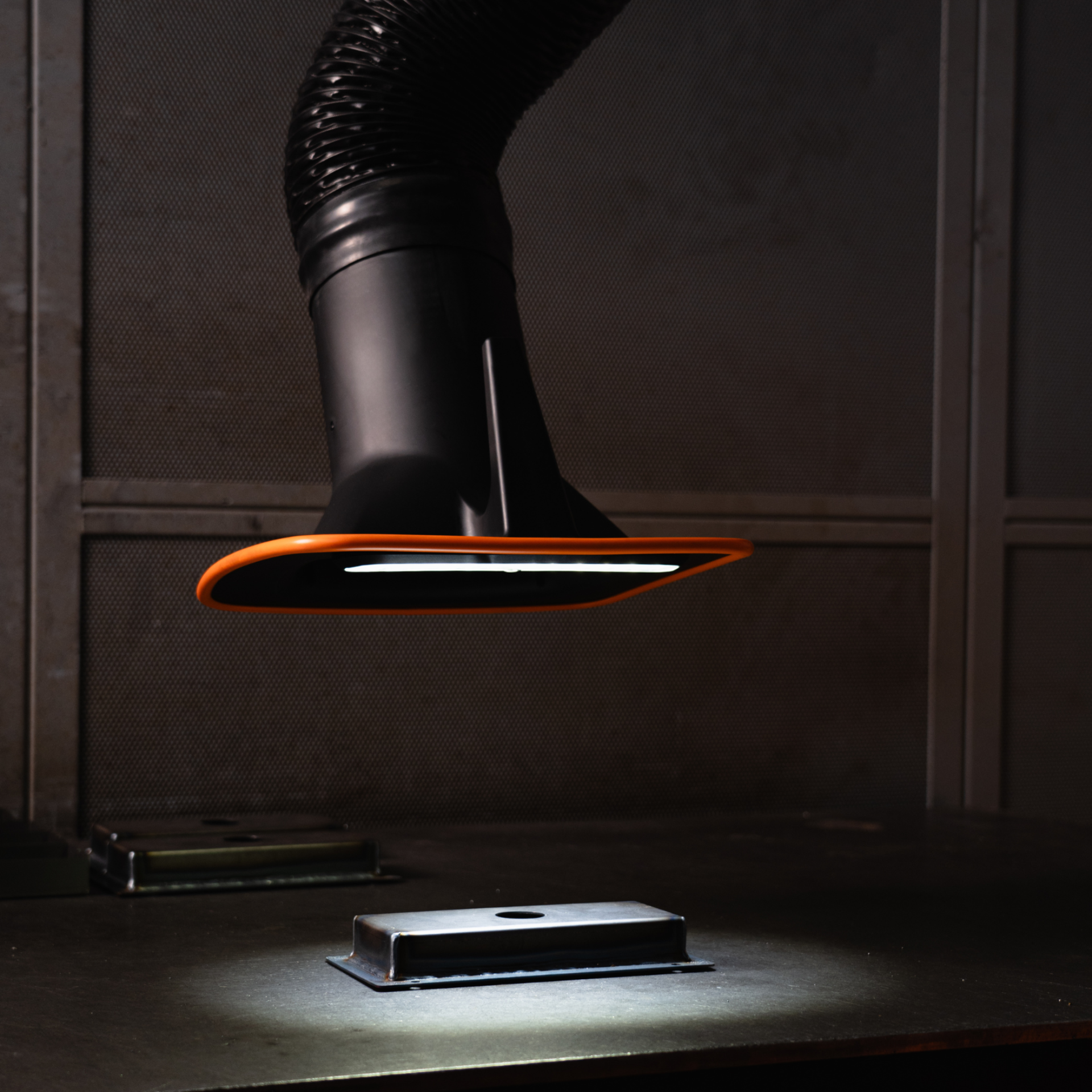 Nahaufnahme eines schwarzen Absaugarms mit orangefarbener Einfassung und integrierter LED-Leuchte, der über einem Metallbauteil auf einem Arbeitstisch positioniert ist.