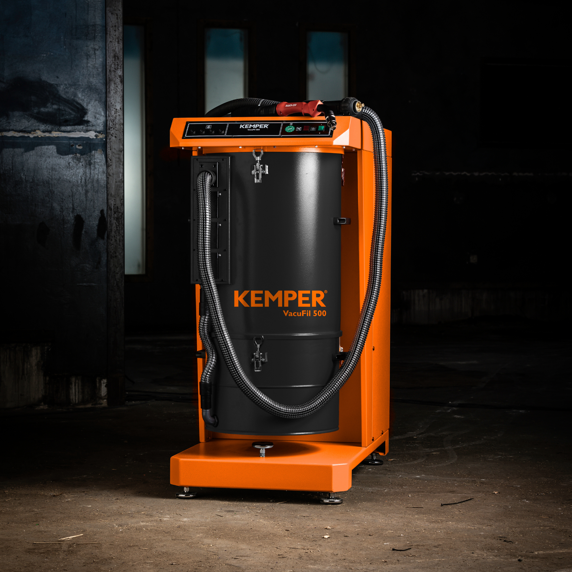 Orange-schwarze KEMPER VacuFil 500 Hochvakuum-Absauganlage mit angeschlossenem Absaugschlauch und Bedienfeld, aufgestellt in einer dunklen Industriehalle mit Betonboden.